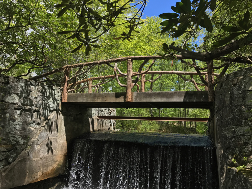 Bridge over a waterfall built in the rustic style using logs and branches titled the Milltown Railings