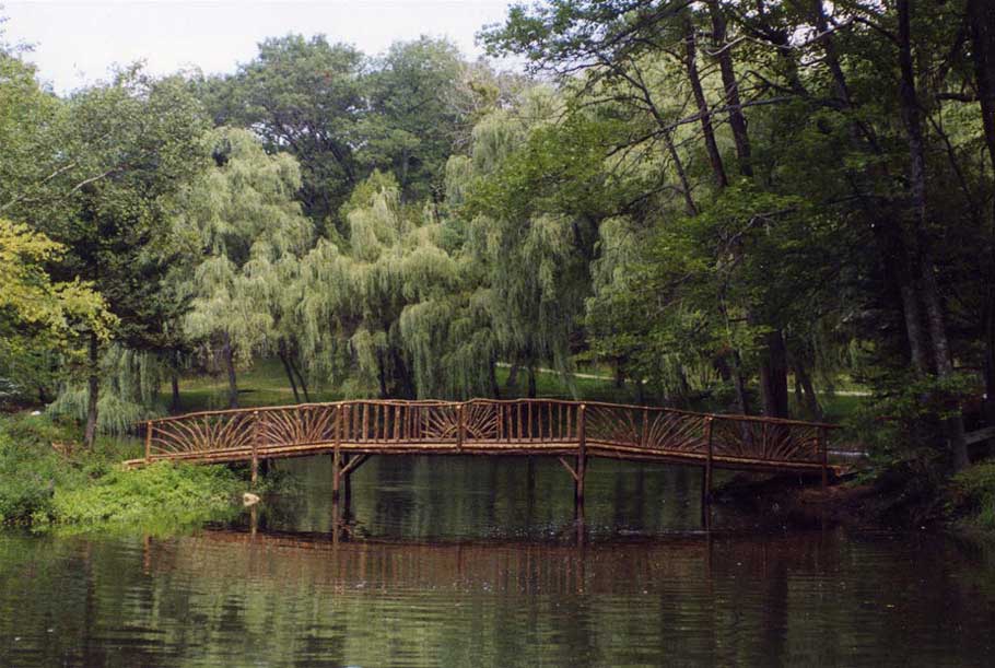 A large rustic bridge crossing pond built using bark-on trees and branches titled the Fisher Bridge