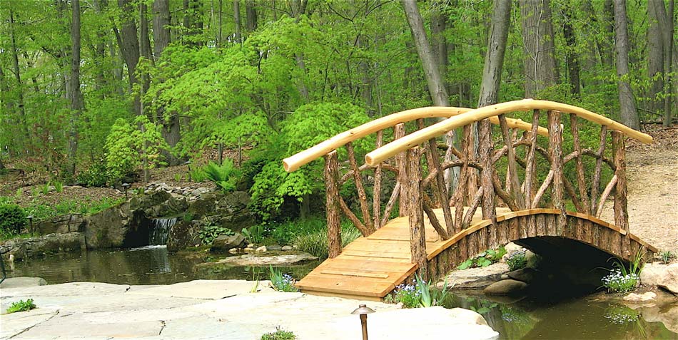 Rustic bridge on a beautifully landscaped yard built using bark-on trees and branches titled Old Westbury Bridge