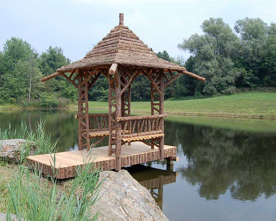 Rustic sitting shelter on the end of a dock built using bark-on trees and branches titled the Van Wormer Shelter