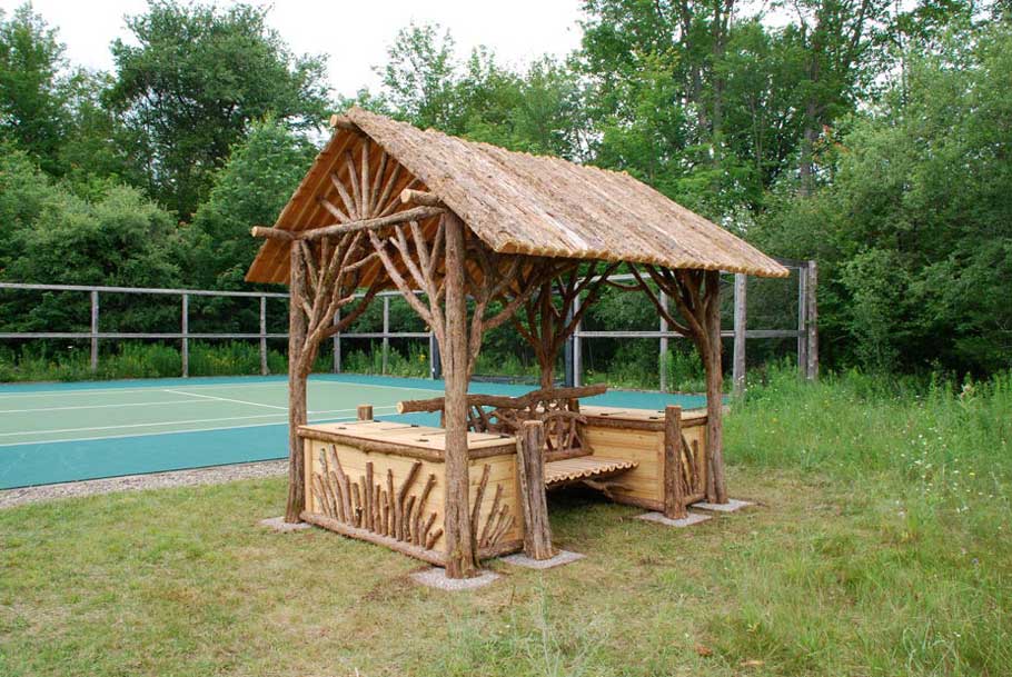 Natural wood covered shelter built with trees and branches titled the Tupper Lake Tennis Shelter